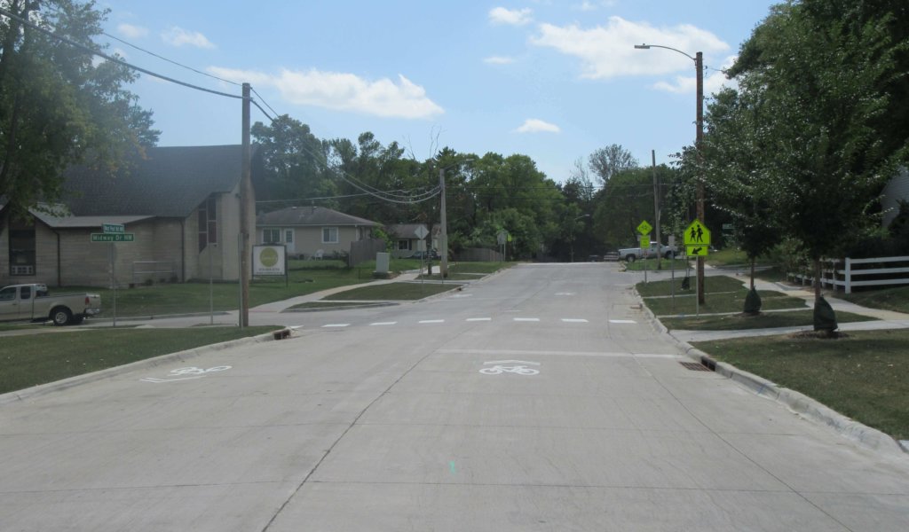 New residential intersection and street pavement with pavement markings, signs, power lines, trees, and vehicle.
