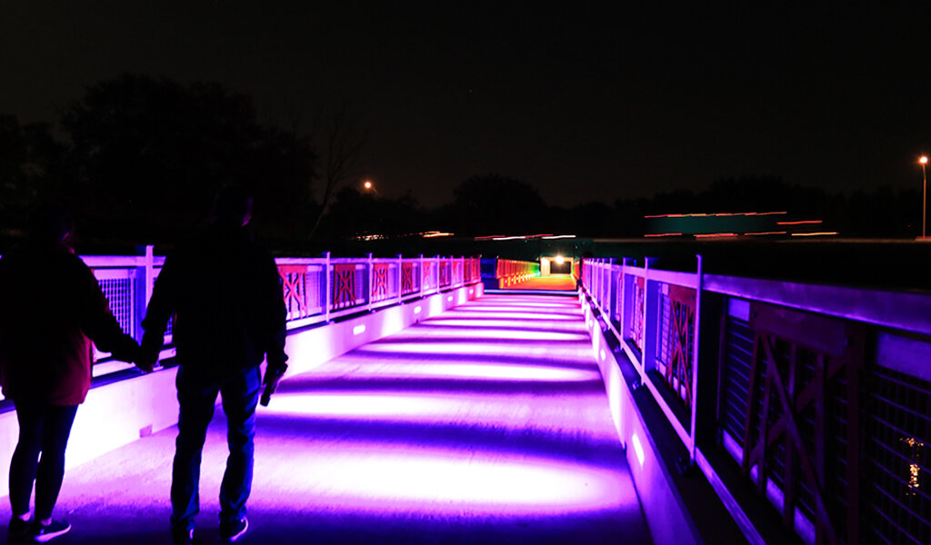 Couple walks across a lit up trail bridge at night