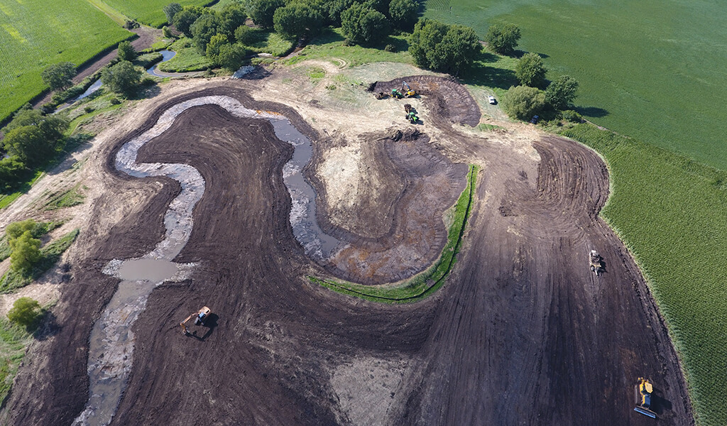 Aerial view of construction at Niebuhr Mitigation bank|update stream with new protections|aerial view of niebuhr mitigation bank|field of reed canary grass|construction machinery creating low water crossing in a stream|snyder & associates employees walking through mitigation bank|construction machinery digging out dirt for stream