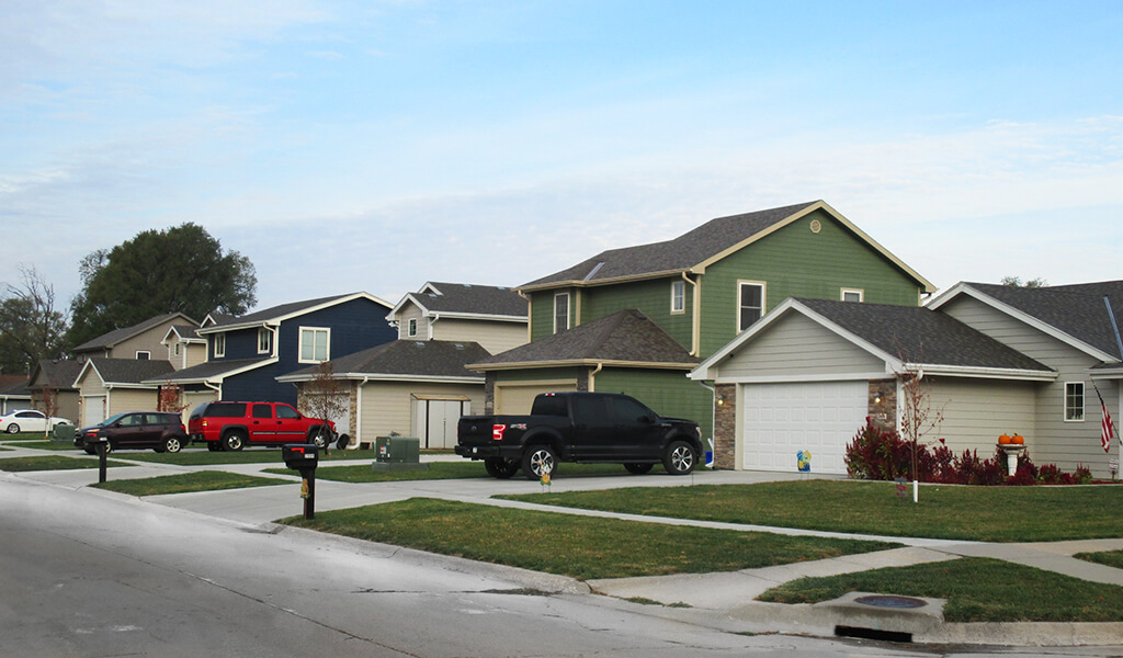 houses on a neighborhood street taken from an angle shot with vehicles in the driveways|Brand new housing on Walnut Grove in Council Bluffs Iowa|Old school surrounded by green grass and native plantings|Flat grass land with trees primed for housing construction to begin|Walnut Grove School prior to demolition|two story square blue house with stone work and green lawn