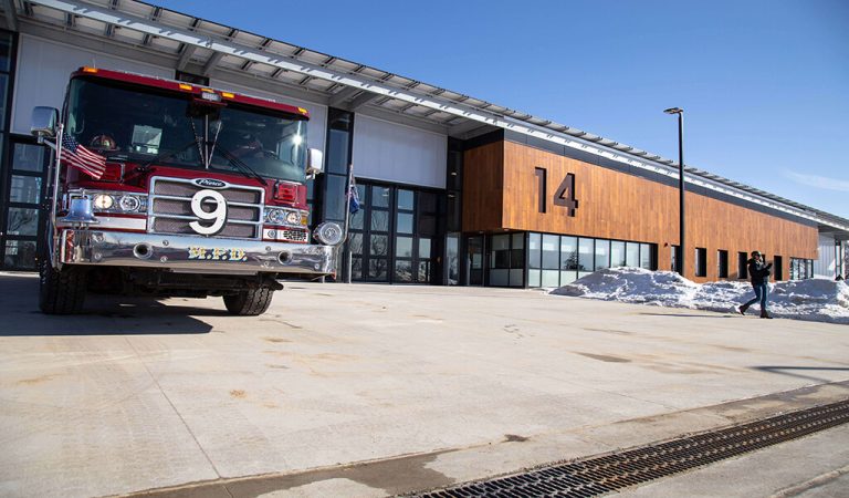 ||Design team stands in-front of red wall decorated with fire fighter items|Newly renovated front of Station 14|fire chief holding a fire hose at ceremonial opening|Cars parked on permeable paver lot in front of Fire Station 14 in Madison.|||