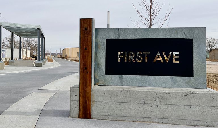 Thick metal signage with large text|Bicyclist ride past the Council Bluffs fire station|Before construction on First Ave|Bicyclist happily wave at on down the bike trail|First Ave Trail in Council Bluffs|Asphalt trail along high school athletic facilities|Council Bluffs bike trail