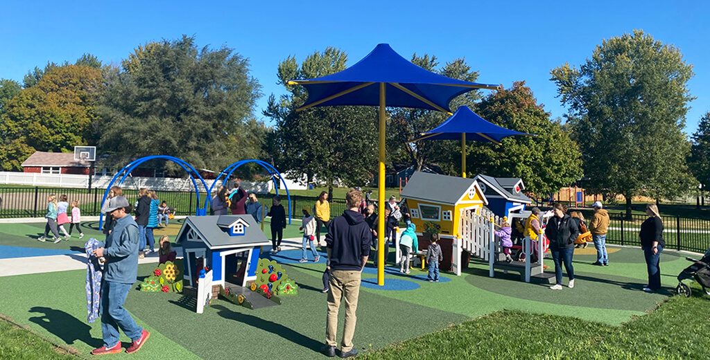 Children of Pella enjoying playing on the Kiwanis Playground|Kids playing on the ADA playground equipment at Pella's Kiwanis Park|The community enjoying playing at the inclusive Kiwanis playground|Kiwanis Playground turf surfacing|Kiwanis Slide and turf ground|Two adults read a playground sign of the list of major donors at Kiwanis Park|ADA Playground equipment at Kiwanis Park in Pella Iowa|Kiwanis Park Bathroom and drinking fountain|Construction and concrete progress at the Kiwanis Park|Phasing Plan for Kiwanis Park Improvements