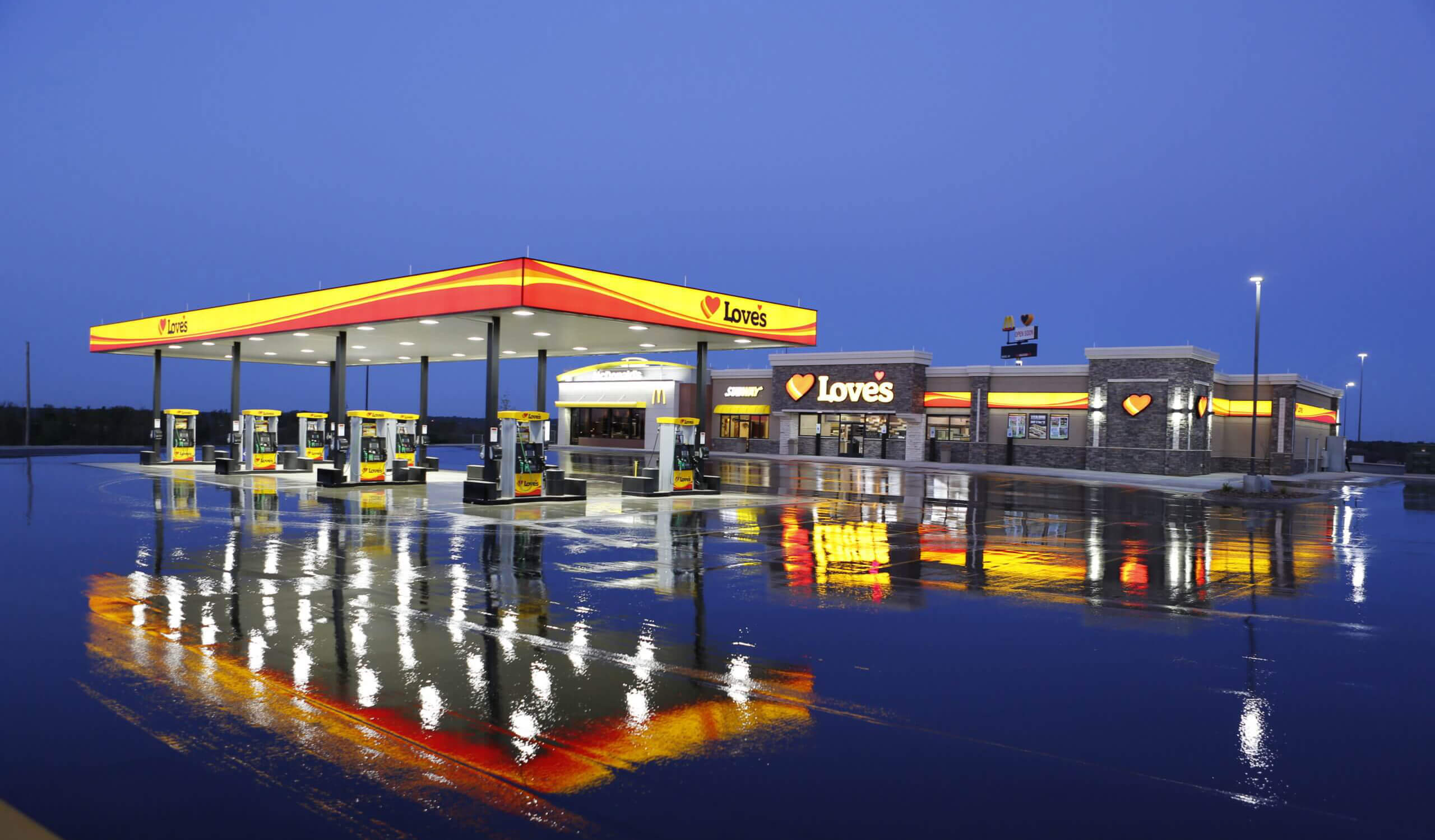 Photo of a Loves truck stop at night on a rainy day|Photo of a Loves truck stop at night on a sunny day|Gas station utilities being constructed.|Construction workers paving cement at a Loves truck stop.