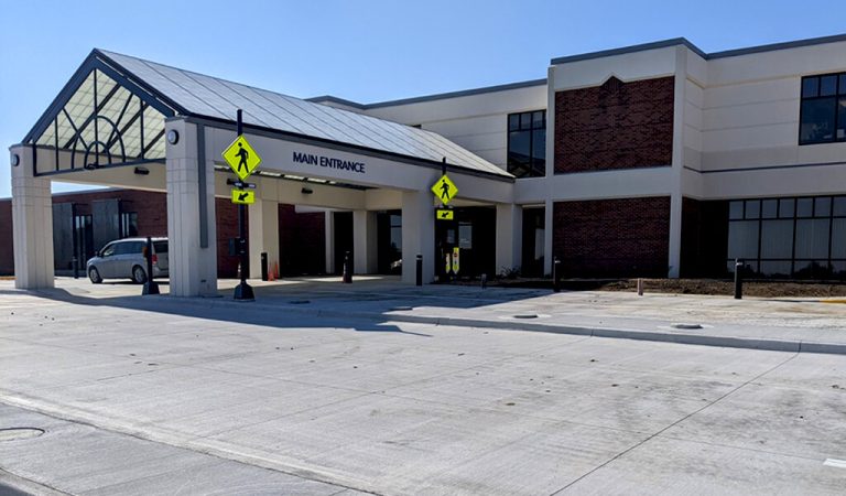 Hospital main entrance|new concrete leading to the main entrance|new parking with cars filling some of the spots|a new dialysis center along a roadway|large newly paved parking lot|Modern health care facility