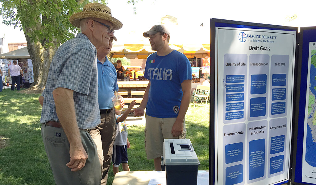 a young man talks with an old man at an outdoor engagement meeting|large brown and green welcome sign at the entrance to large park|two men shovel wet concrete into place|concrete roadway with shattered concrete in middle of drive|large brown welcome sign at entrance of park