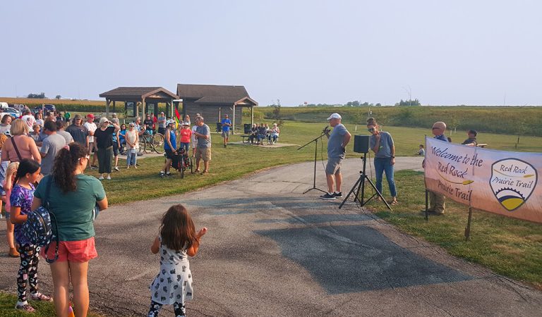 |Prairie City Welcome signage Town Square|Large green plants and trees on either side of a bike trail|Winding trail heads towards a Casey's Gas station|Child bikes a trail through a small town in Iowa|Small town welcome sign with a trail and road next to it|Prairie City town square||