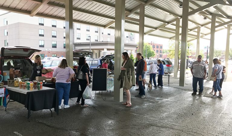 people gathered around tables under metal pavilion||large brick and cement sign for farmers market|people attending farmers market|Group of people prepare to cut red ribbon|metal pavilion housing farmers market|