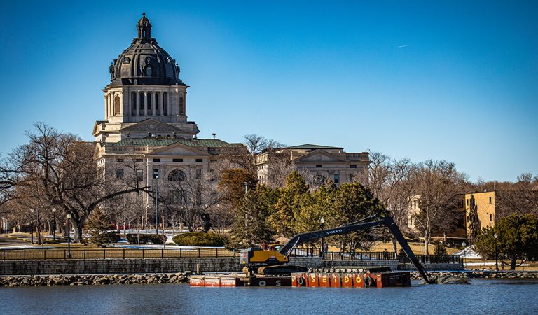 South Dakotas capitol building behind dredging equipment on lake|excavator on floating platform|excavator dredging from shoreline|sediment being pulled from lake bed|aerial view of capitol lake in south dakota|Dump truck being filled with sediment from of South Dakota's Capitol Lake