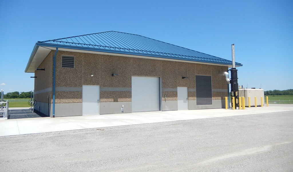 cement building with blue roof|grey wall of electrical pannels|multiple pumps in cement room|three oval motors in a row|looking down on multi-level facility floors|