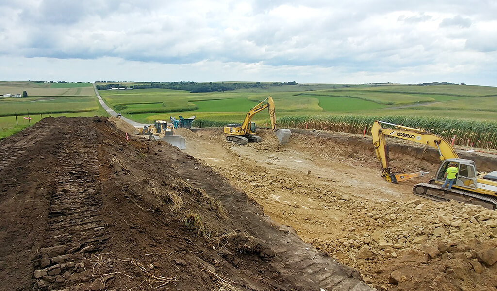 Large construction equipment moves dirt|yellow speed sign gives 35 mph limit|uphill road|area dug out in preparation of road|new asphalt laid on road|white GIS map shows roadway plotting|man in safety vest standing beside road counting traffic