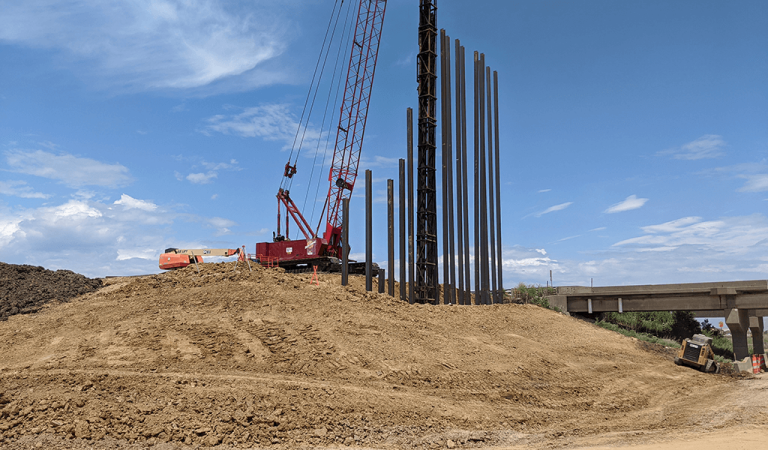 red crane with large metal beams standing straight up|crane building interstate overpass|dirt construction site with work going on in the background||view from under overpass looking at beams