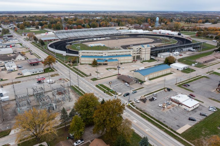Aerial view of a dirt oval race track with surrounding grandstands and a multi-level museum building labeled National Sprint Car Hall of Fame & Museum, plus parking lots and fall-colored trees in the background.