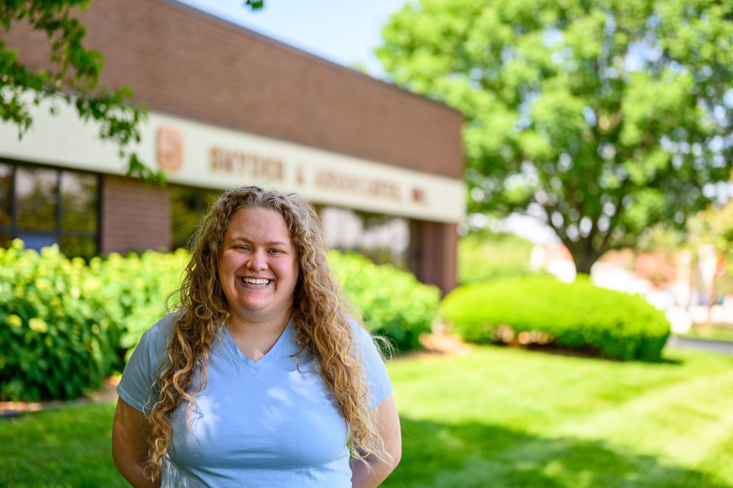 Smiling woman with curly hair in light blue scrubs outdoors, sunny day with green plants and a brick building in the background