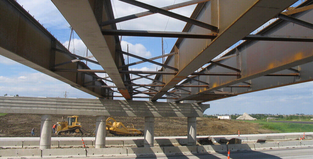Bridges Under a large steel bridge frame at a road construction site with a bulldozer and excavator on a dirt embankment and concrete barriers nearby.