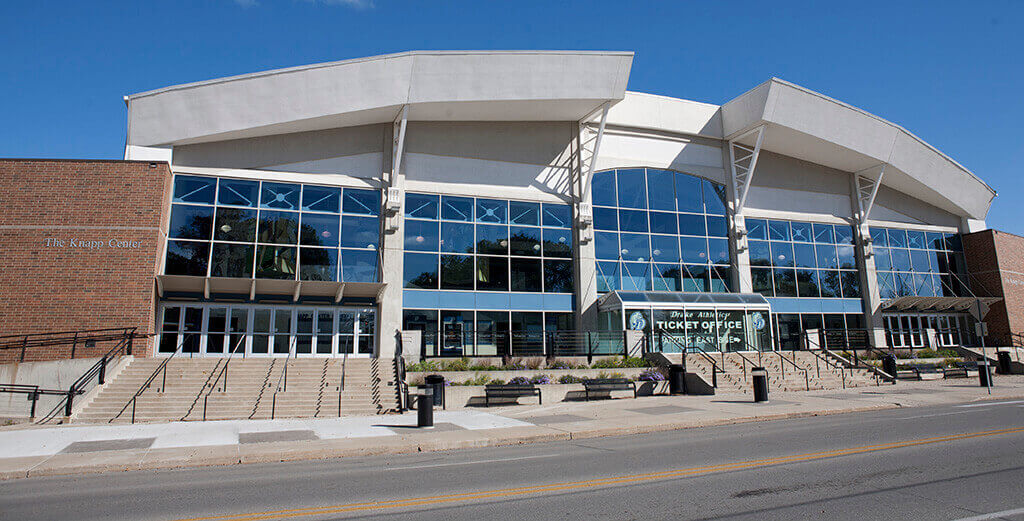 Front entrance of a large arena with glass walls, ticket office, and wide concrete stairs under a blue sky.