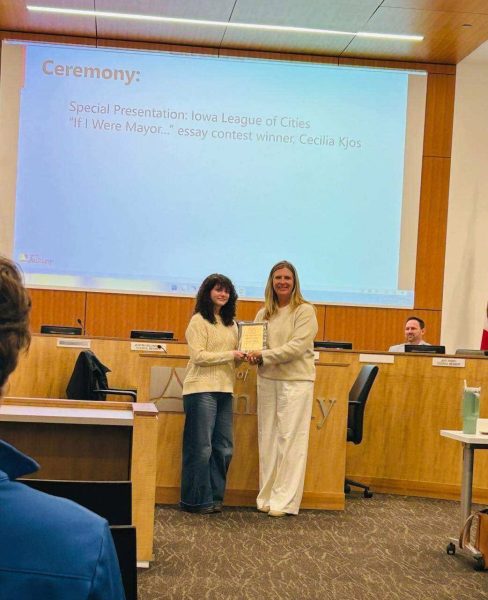 Two women stand at a podium area exchanging a plaque during a ceremony in a conference room with a large screen behind them.