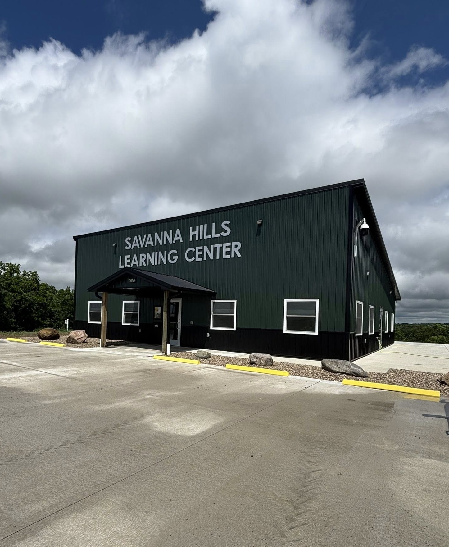Green school building with 'Savanna Hills Learning Center' sign, front entrance canopy, parking lot, and cloudy sky above.