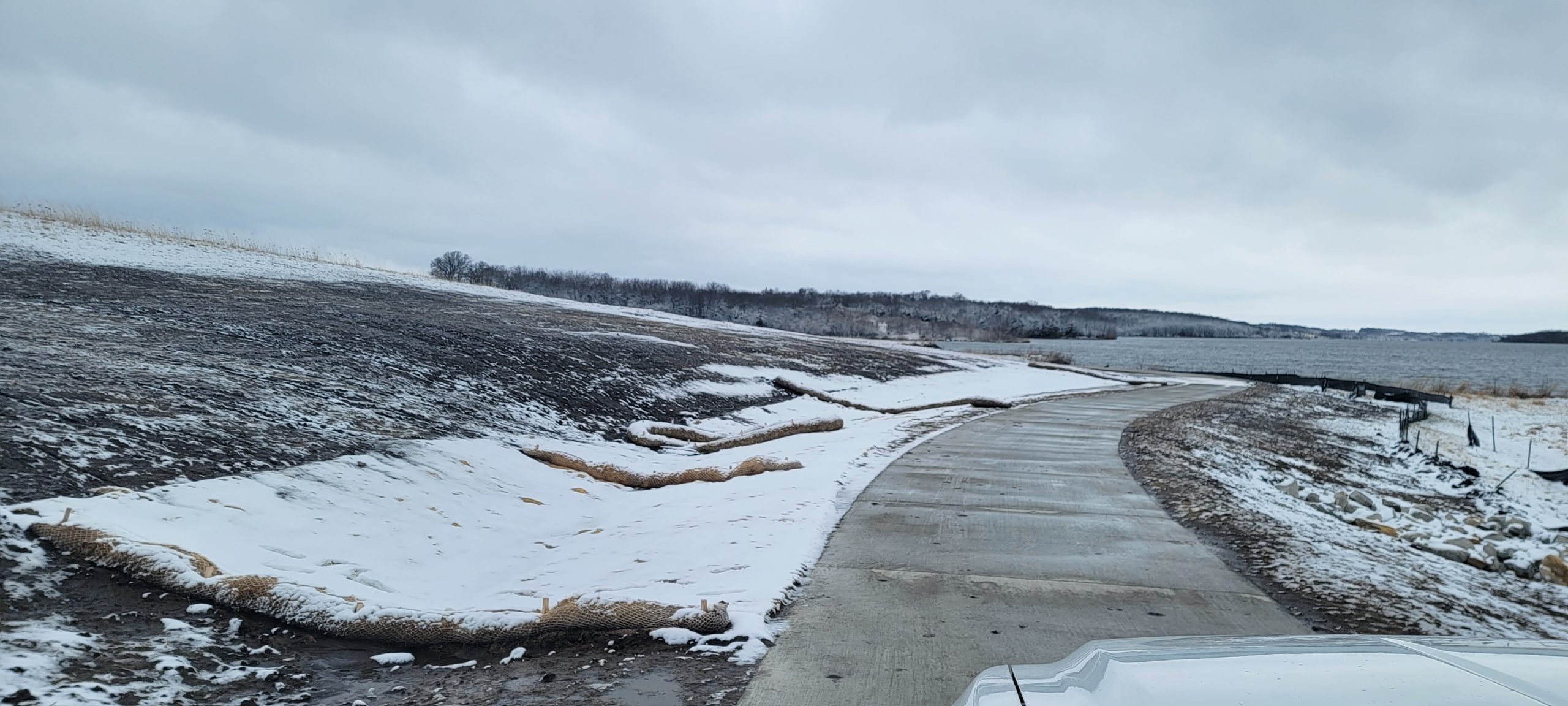 Snowy lakeshore with a curved concrete path, overcast sky, and a distant tree line along the water edge.