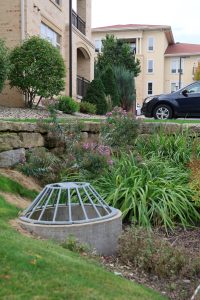 Circular metal storm drain cover in a rock-filled garden bed beside beige apartment buildings; a blue car is parked nearby.