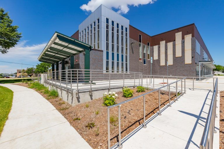 Exterior view of a modern brick and glass building with a ramp and metal railings under a bright blue sky, sunny day.