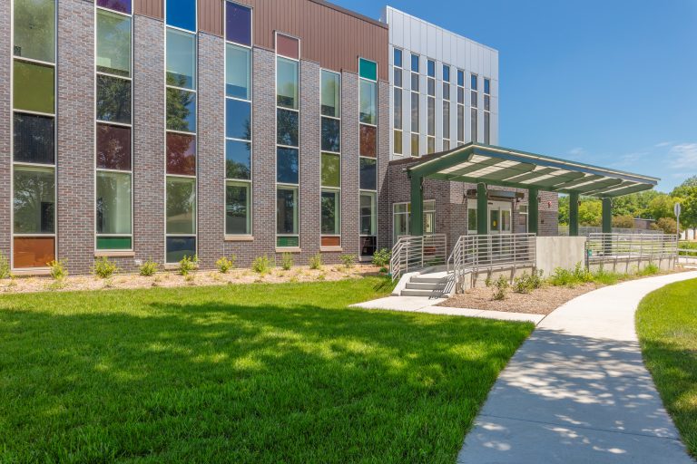 Front view of a modern brick building with tall, colorful windows and a green-framed entrance under a glass canopy, adjacent to a curved walkway and manicured lawn.