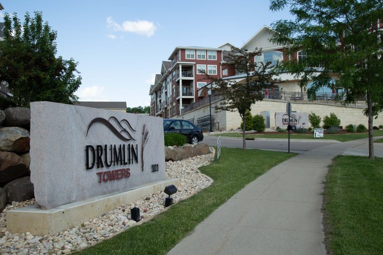 Exterior view of a residential complex with a large stone sign reading 'DRUMLIN TOWERS' and modern brick buildings in the background under a blue sky