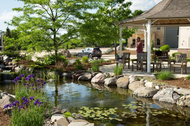 Man standing on a covered patio beside a landscaped backyard with a pond, rocks, and purple irises.