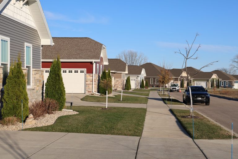 Suburban street with rows of attached garages, trimmed lawns, and a black car parked on the right against a blue sky.
