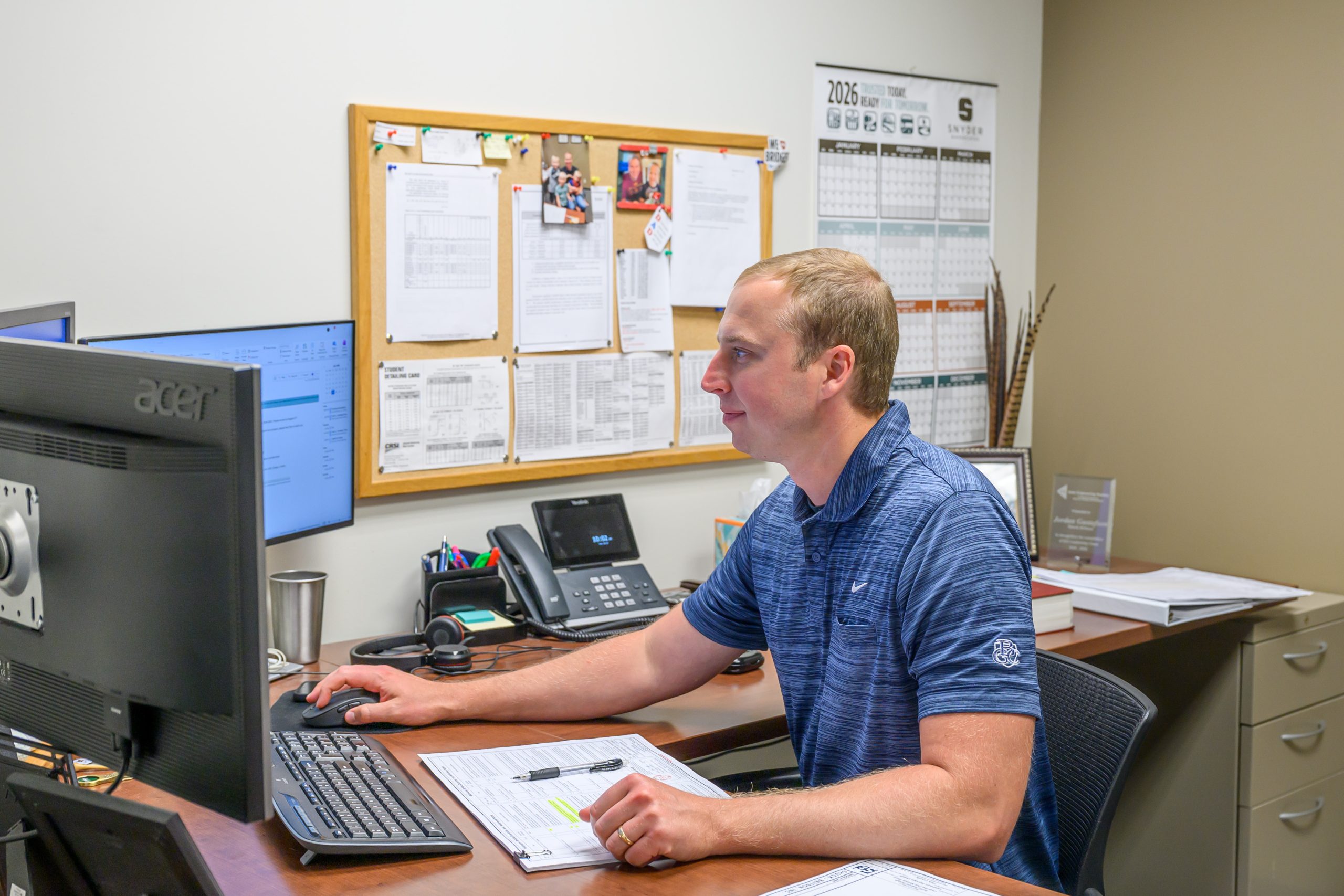 2026_EmployeeSpotlight_JordanGustafson_020 Man at a wooden desk typing on a keyboard with two computer monitors in an office setting, surrounded by papers and office supplies.